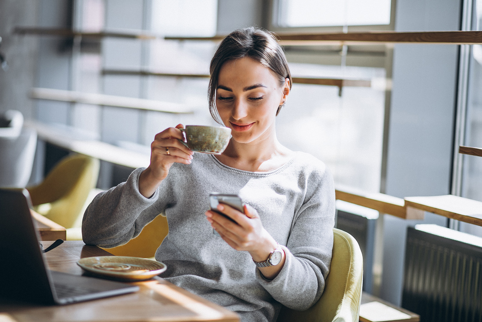 Junge Frau sitzend am Tisch mit Kaffeetasse, Handy und Laptop vor sich